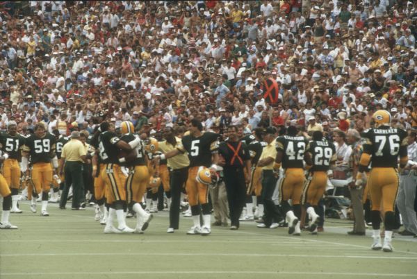 View of the Packers sideline after a play with Gary Ellerson (#42), Ken Stills (#29) and Alphonso Carreker (#76). The game against the New York Jets was preseason and took place at Camp Randall Stadium. This was the largest crowd ever to see the Packers play in Wisconsin and the first pro football game ever held in Madison. The Packers won the game 38 to 14.