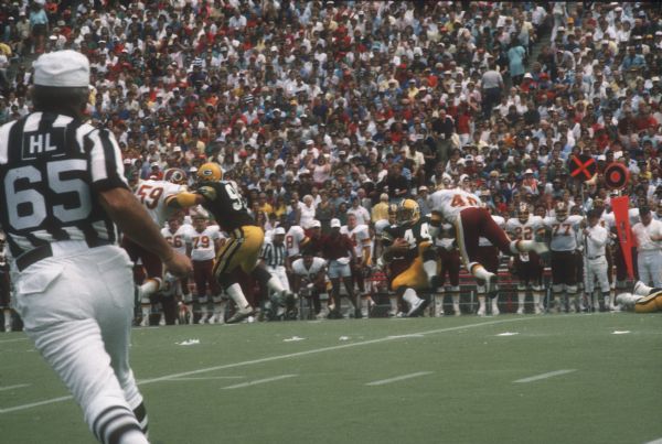 A Green Bay Packer (most likely) Von Mansfield (#44) takes a hit during a preseason game against the Washington Redskins that was played at Camp Randall Stadium in Madison, WI. A referee is visible in the left foreground. The Packers lost 0 to 33.