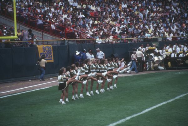 Elevated view of a line of Green Bay Packers cheerleaders dancing in the end zone. A band is visible in the background on the sideline. The Packers' cheerleaders were known as the Sideliners from 1977-1986.