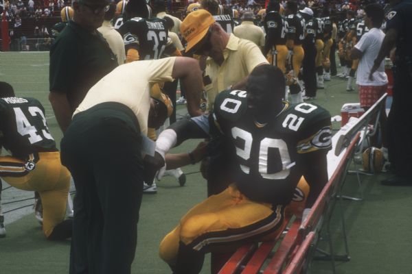 Green Bay Packer running back Kelly Cook (#20) sits on a bench getting some assistance on the sideline during a preseason game against the Washington Redskins that took place at Camp Randall Stadium. Packers Larry Morris (#43) and John Simmons (#32) are in the background. The Packers lost 0 to 33.