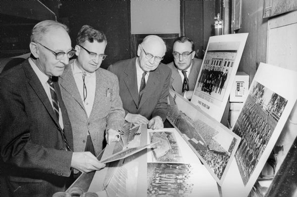 Four men examining various Packers memorabilia, including images of the first Packers team from 1919, the team from 1929, other unidentified team lineups, as well as Green Bay stadiums through the years.