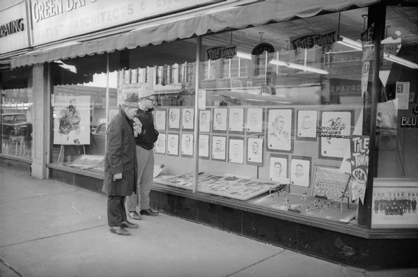 Two teenage boys stand on the sidewalk looking at an art store window display featuring illustrations of the Packer players. The Packers were about to play for, and would go on to win, their seventh title as World Champions of Pro Football. A large portrait of head coach Vince Lombardi can be seen behind the teens.
