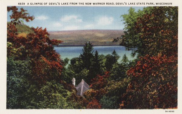Colorized postcard of Devil's Lake and bluffs seen through the trees. Some of the trees have autumn color. A roof and chimney are visible. The text at the top reads: "A Glimpse Of Devil's Lake From The New Warner Road, Devil's Lake State Park, Wisconsin."