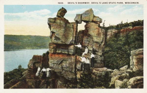 Colorized postcard of the rock formation called the Devil's Doorway in Devil's Lake State Park. Trees can be seen to the right and below. The lake, bluffs and sky are visible in the background. Sitting and standing around the formation are five women and three men. They are wearing white shirts and dark pants or skirts. The text above reads: "Devil's Doorway, Devil's Lake State Park, Wisconsin."