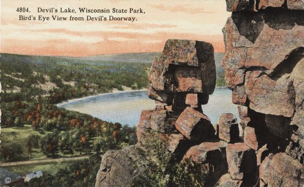 Colorized postcard of the rock formation called the Devil's Doorway in Devil's Lake State Park. Trees can be seen below. The lake, bluffs, roads and buildings are visible to the left. The sky is orange. The text above reads: "Devil's Lake, Wisconsin State Park, Bird's Eye View from Devil's Doorway."