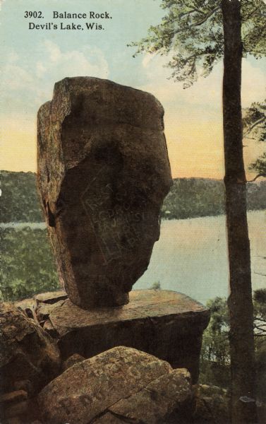 Colorized postcard of Balance Rock, a formation in Devil's Lake State Park. A tree is on the right and the lake, trees, bluffs and sky are in the background. There are carved initials and other markings on the rock formation. The text at the top reads: "Balance Rock, Devil's Lake, Wis."
