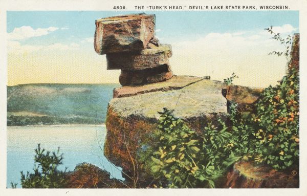 Colorized postcard of the Turk's Head, a formation in Devil's Lake State Park. Foliage is in the foreground. The lake, trees, bluffs and sky are in the background. The text at the top reads: "The 'Turk's Head,' Devil's Lake State Park, Wisconsin."