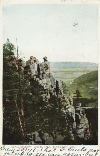 Colorized postcard of the rock formation called the Devil's Doorway in Devil's Lake State Park. Trees are on the left and below. Bluffs, trees, sky and a road are in the background. Caption reads: "Devils [sic] Doorway, Looking East, Devils [sic] Lake, Wis., Wis."