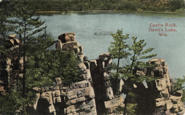 Colorized postcard of the rock formation called Castle Rock in Devil's Lake State Park. Trees can be seen throughout. The lake, far shoreline, bluffs and more trees are visible in the background. The text in the upper right corner reads "Castle Rock, Devil's Lake, Wis."