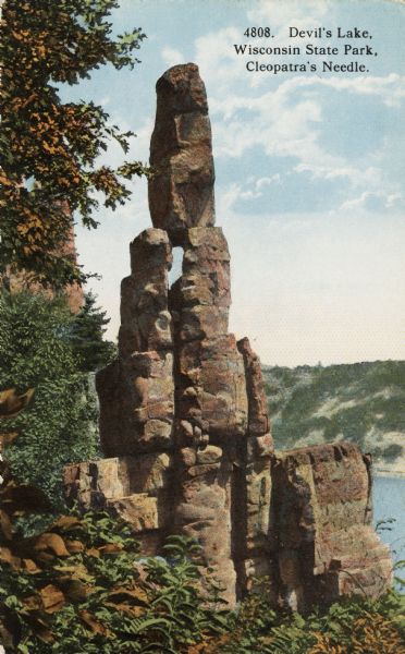 Colorized postcard of the rock formation called Cleopatra's Needle in Devil's Lake State Park. Trees and foliage can be seen to the left and below. The lake, bluffs and sky are visible in the background. The text above reads "Devil's Lake, Wisconsin State Park, Cleopatra's Needle."