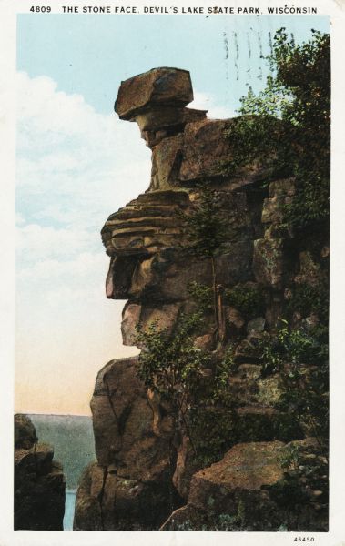 Colorized postcard of the rock formation called the Stone Face in Devil's Lake State Park. Trees and foliage can be seen to the right and below. The lake, bluffs and sky are visible in the background. The text above reads "The Stone Face, Devil's Lake State Park, Wisconsin."