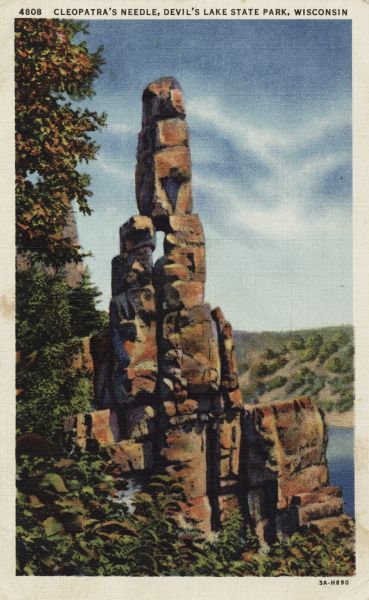 Colorized postcard of the rock formation called Cleopatra's Needle in Devil's Lake State Park. Trees can be seen to the left and below. The lake, bluffs and sky are visible in the background. The text above reads "Cleopatra's Needle, Devil's Lake State Park, Wisconsin."