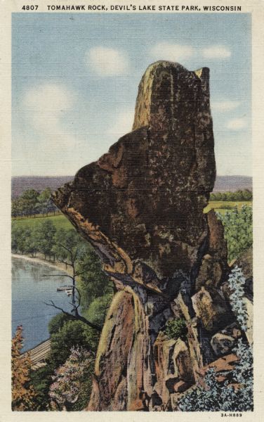 Colorized postcard of the rock formation called Tomahawk Rock in Devil's Lake State Park. Foliage can be seen below. The lake, bluffs, trees and sky are visible in the background. The text above reads "Tomahawk Rock, Devil's Lake State Park, Wisconsin."