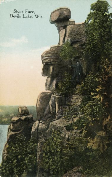 Colorized postcard of the rock formation called the Stone Face in Devil's Lake State Park. Trees and shrubs can be seen all around. A man is standing to the right of the "face." The lake, bluffs and sky are visible in the background. The text above reads "Stone Face, Devil's Lake, Wis."