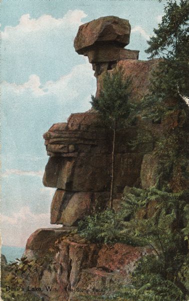 Colorized postcard of the rock formation called the Stone Face in Devil's Lake State Park. Trees and shrubs can be seen all around. The sky is visible in the background. The text above reads "Devil's Lake, Wis. The Stone Face."