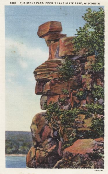 Colorized postcard of the rock formation called the Stone Face in Devil's Lake State Park. Trees and shrubs can be seen all around. The lake, bluffs and sky are visible in the background. The text above reads "The Stone Face, Devil's Lake State Park, Wisconsin."