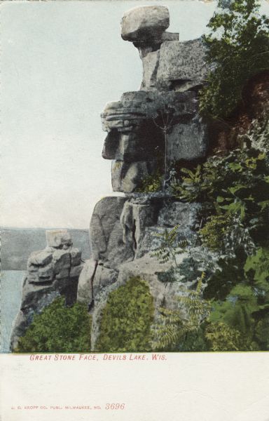 Colorized postcard of the rock formation called the Great Stone Face in Devil's Lake State Park. Trees and shrubs can be seen all around. The lake, bluffs and sky are visible in the background. The text above reads "Great Stone Face, Devil's Lake, Wis."