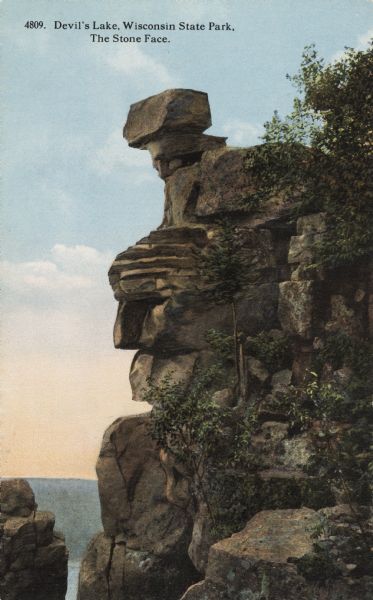 Colorized postcard of the rock formation called the Stone Face in Devil's Lake State Park. Trees and shrubs can be seen all around. The lake, bluffs and sky are visible in the background. The text above reads "Devil's Lake, Wisconsin State Park, The Stone Face."