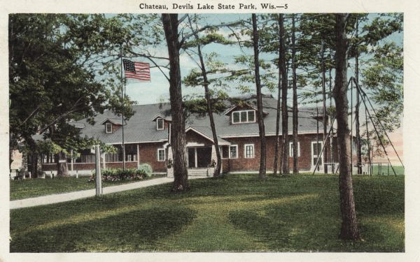 Colorized postcard of the Chateau in Devil's Lake State Park. Trees, shrubs, flowers and grass surround the building. An American flag flies in front and a swing set is on the right. The text above reads "Chateau, Devil's Lake State Park, Wis."