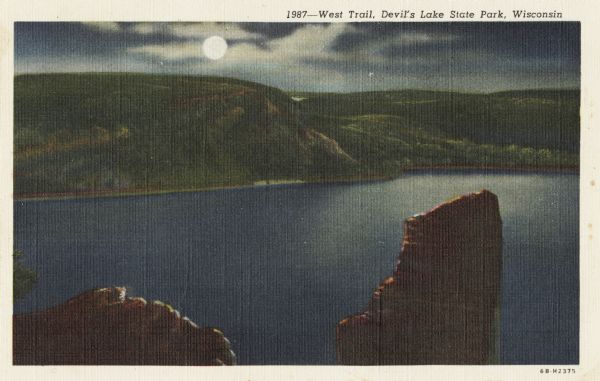 Colorized postcard of Devil's Lake by moonlight. Two rock formations are in the foreground. The lake, trees, bluffs and sky are in the background. The text above reads: "West Trail, Devil's Lake State Park, Wisconsin."
