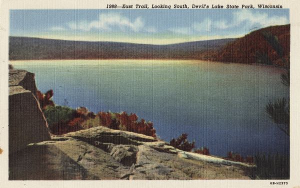 Colorized postcard of Devil's Lake, looking south from the East Trail. Rock formations are in the lower left, with trees below. Bluffs, trees and sky are in the background. The text above reads: "East Trail, Looking South, Devil's Lake State Park, Wisconsin."