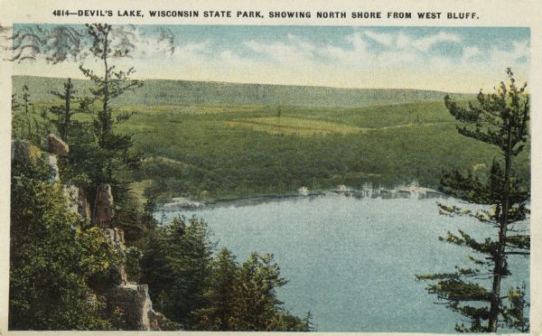 Colorized postcard of Devil's Lake. Rock formations are on the left side. Bluffs and sky are in the background. There are buildings along the shoreline. The text above reads: "Devil's Lake, Wisconsin State Park, Showing North Shore From East Bluff."
