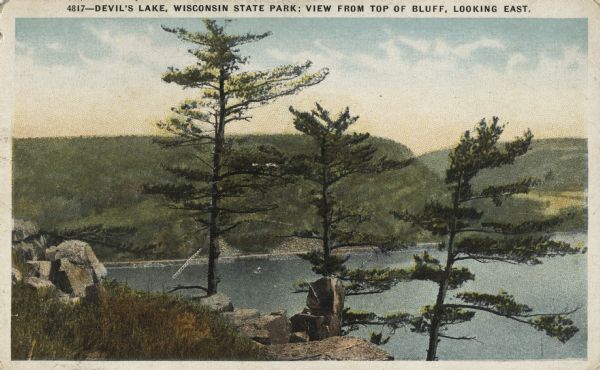 Colorized postcard of Devil's Lake. Rock formations are in the foreground. Trees can be seen throughout. Bluffs and sky are visible in the background. The text above reads "Devil's Lake, Wisconsin State Park; View From the Top of Bluff, Looking East."
