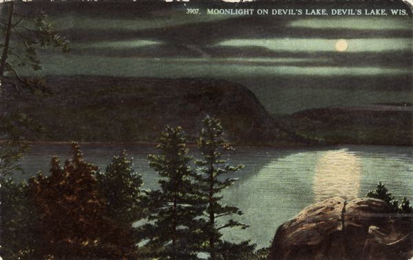 Colorized postcard of Devil's Lake by moonlight. A rock formation and trees are in the foreground. The lake, trees, bluffs and sky are in the background. The text above reads: "Moonlight On Devil's Lake, Devil's Lake, Wis."