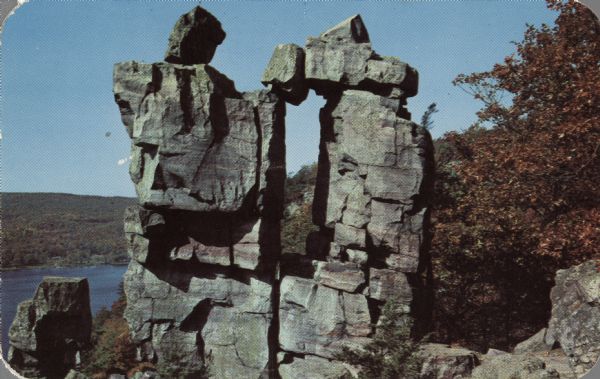 Color postcard view of the rock formation called the Devil's Doorway in Devil's Lake State Park. Trees are changing color. The lake, bluffs and sky are in the background.