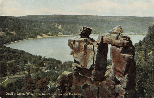 Colorized postcard view of the rock formation called the Devil's Doorway in Devil's Lake State Park. Trees are growing among the rocks. The lake, bluffs, buildings, roads and sky are in the background. There are carved initials and other markings on the rock formation. The text below reads: "Devil's Lake, Wis. The Devil's Doorway and the Lake."