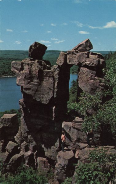 Color postcard view looking down at the rock formation called the Devil's Doorway in Devil's Lake State Park. The lake, bluffs and sky are in the background. Two people are standing on the rocks. The text above reads: "Devil's Doorway in Devil's Lake State Park."