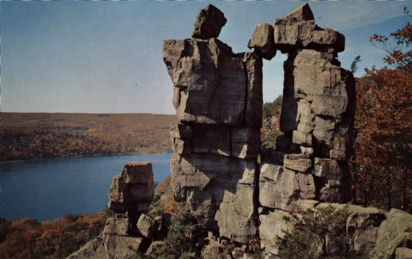 Color postcard view looking down at the rock formation called the Devil's Doorway in Devil's Lake State Park. The trees are changing color. The lake, bluffs and sky are in the background.