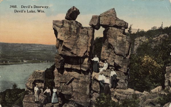 Colorized postcard of the rock formation called the Devil's Doorway in Devil's Lake State Park. Trees can be seen to the right and below. The lake, bluffs and sky are visible in the background. Sitting and standing around the formation are five women and three men. They are wearing light shirts and dark pants or skirts. There are carved initials and other markings on the rock formation. The text above reads "Devil's Doorway, Devil's Lake, Wis."