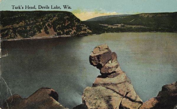 Colorized postcard of the rock formation called the Turk's Head in Devil's Lake State Park. The lake, bluffs, trees and sky are visible in the background. The text above reads "Turk's Head, Devil's Lake, Wis."