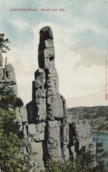 Colorized postcard of the rock formation called Cleopatra's Needle in Devil's Lake State Park. Trees and foliage are on the left and below. The lake, bluffs and sky are in the background. Caption reads: "Cleopatra's Needle, Devils [sic] Lake, Wis."