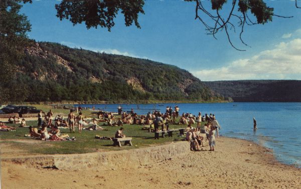 Color postcard of the bathing beach at Devil's Lake State Park with a  view of the East Bluff. The lake, bluffs and sky are in the background. Many visitors are on the beach, and a parking lot is off to the left.