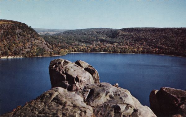Color postcard view of a rock formation above the lake in Devil's Lake State Park. The lake, bluffs, trees and sky are in the background.