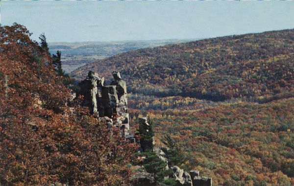 Color postcard of the view from the East Bluff looking towards Lake Wisconsin. The valley is filled with trees that are turning to autumn color. Some rock formations are visible on the left side. In the far distance, Lake Wisconsin can barely be seen.