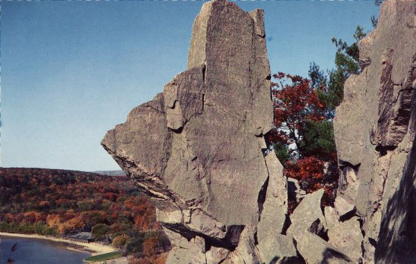 Color postcard view of the rock formation called Tomahawk Rock in Devil's Lake State Park. The lake and beach, trees, bluffs and sky are visible in the background. The trees have changed to fall color. A building is on the beach in the far background.