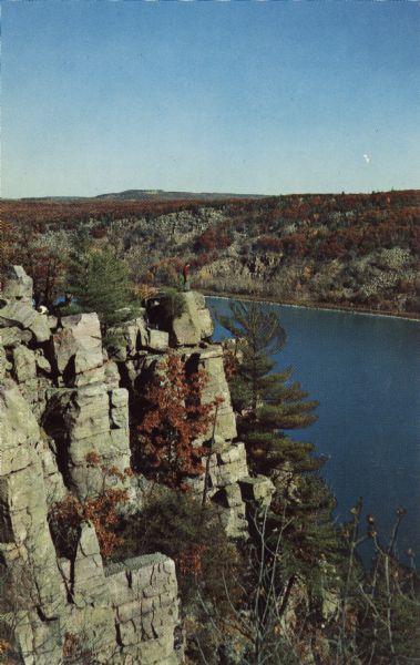 Color postcard of the ramparts of the West Bluff in Devil's Lake State Park. Trees can be seen throughout and have fall color. The lake, bluffs and sky are visible in the background. Several visitors are standing on top of the formation.