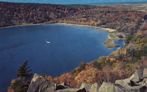 Color postcard of the lake viewed from the East Bluff Trail in Devil's Lake State Park. Trees can be seen all around and it is autumn. Rock formations are in the foreground. A building is on the beach.