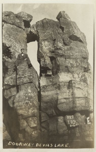 Photographic postcard of the rock formation called the Devil's Doorway in Devil's Lake State Park. A man is sitting on the formation, wearing a suit and a cap. Graffiti is visible on the rocks. Caption reads: "Doorway, Devils [sic] Lake."