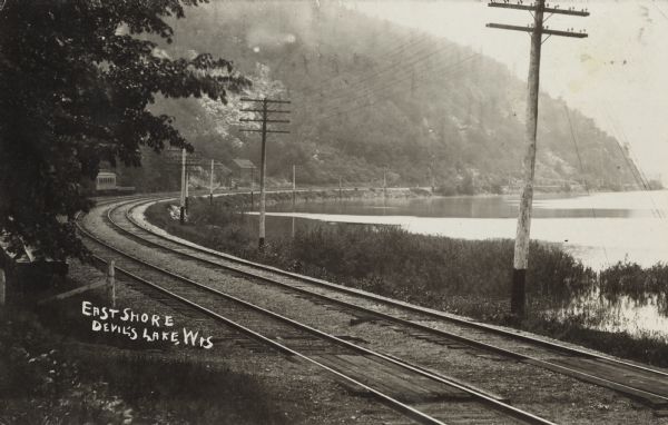 Photographic postcard of the railroad tracks around the East Shore of the lake in Devil's Lake State Park. Telephone poles are between the tracks and the shoreline. Bluffs, buildings and trees are in the background. The text below reads: "East Shore, Devil's Lake, Wis."