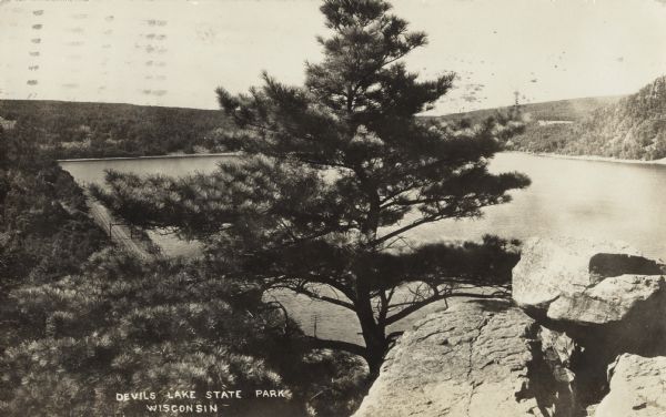 Photographic postcard of rocks and trees above the lake in Devil's Lake State Park. Below on the left railroad tracks run along the shoreline. Trees and bluffs are in the background. The text below reads "Devil's Lake State Park, Wisconsin."