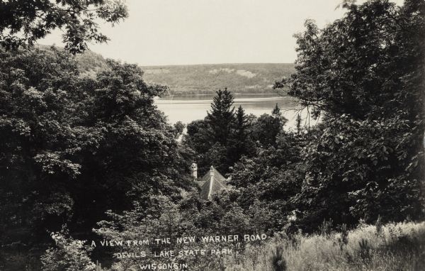 Photographic postcard view from hill of Devil's Lake and bluffs seen through the trees. A roof and chimney are below the hill. The text at the bottom reads: "A View From the New Warner Road, Devil's Lake State Park, Wisconsin."