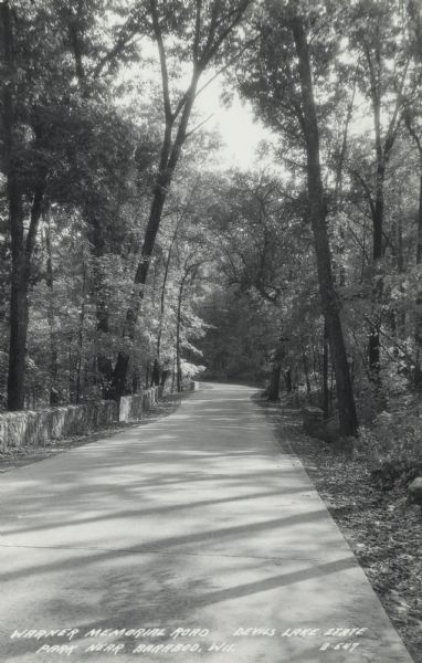 Photographic postcard of the Warner Memorial Road in Devil's Lake State Park. Trees line both sides of the road, and there is a stone wall on the left. The text below reads: "Warner Memorial Road, Devil's Lake State Park Near Baraboo, Wis."