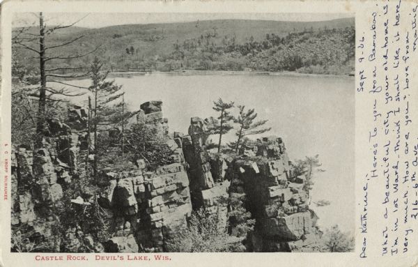 Black and white postcard of the rock formation called Castle Rock in Devil's Lake State Park. Trees can be seen throughout. The lake, bluffs and sky are visible in the background. The text below reads: "Castle Rock, Devil's Lake, Wis."