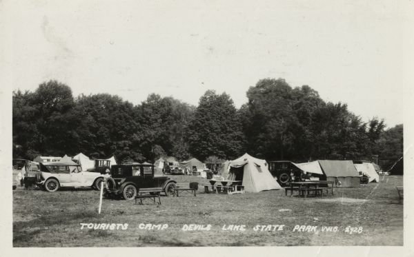 Photographic postcard of a tourist camp at Devil's Lake State Park. A scene of picnic tables, automobiles and tents pitched among trees and grass. Text at the foot reads "Tourist Camp, Devil's Lake State Park, Wis."