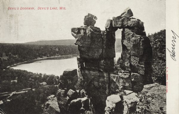 Black and white postcard of the rock formation called the Devil's Doorway in Devil's Lake State Park. Graffiti is visible on the formation. The lake, bluffs, roads and sky can be seen in the background. The text above reads: "Devil's Doorway, Devil's Lake, Wis."