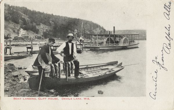Black and white postcard of the boat landing in front of Cliff House, a resort hotel, in Devil's Lake State Park. Two men show off their catch of seven fish while standing in a rowboat pulled up on the shore. A steamboat and several row boats are moored to the pier, and standing at the end are two visitors. Bluffs, buildings and trees are visible in the background. Caption reads: "Boat Landing, Cliff House, Devil's Lake, Wis."